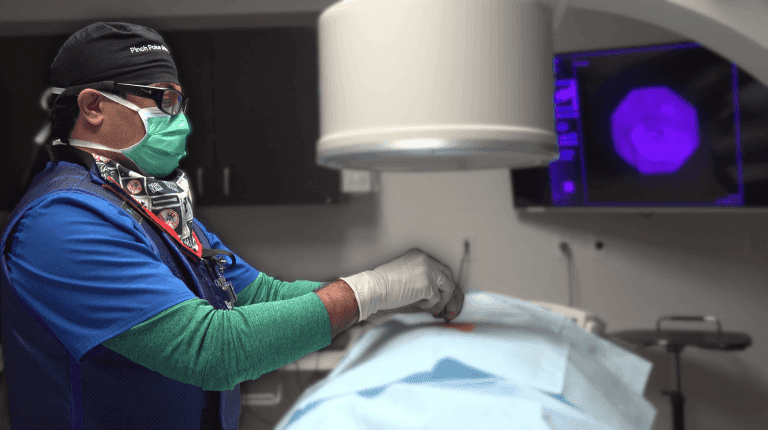 Surgeon in scrubs using medical equipment during a procedure, with a monitor in the background.