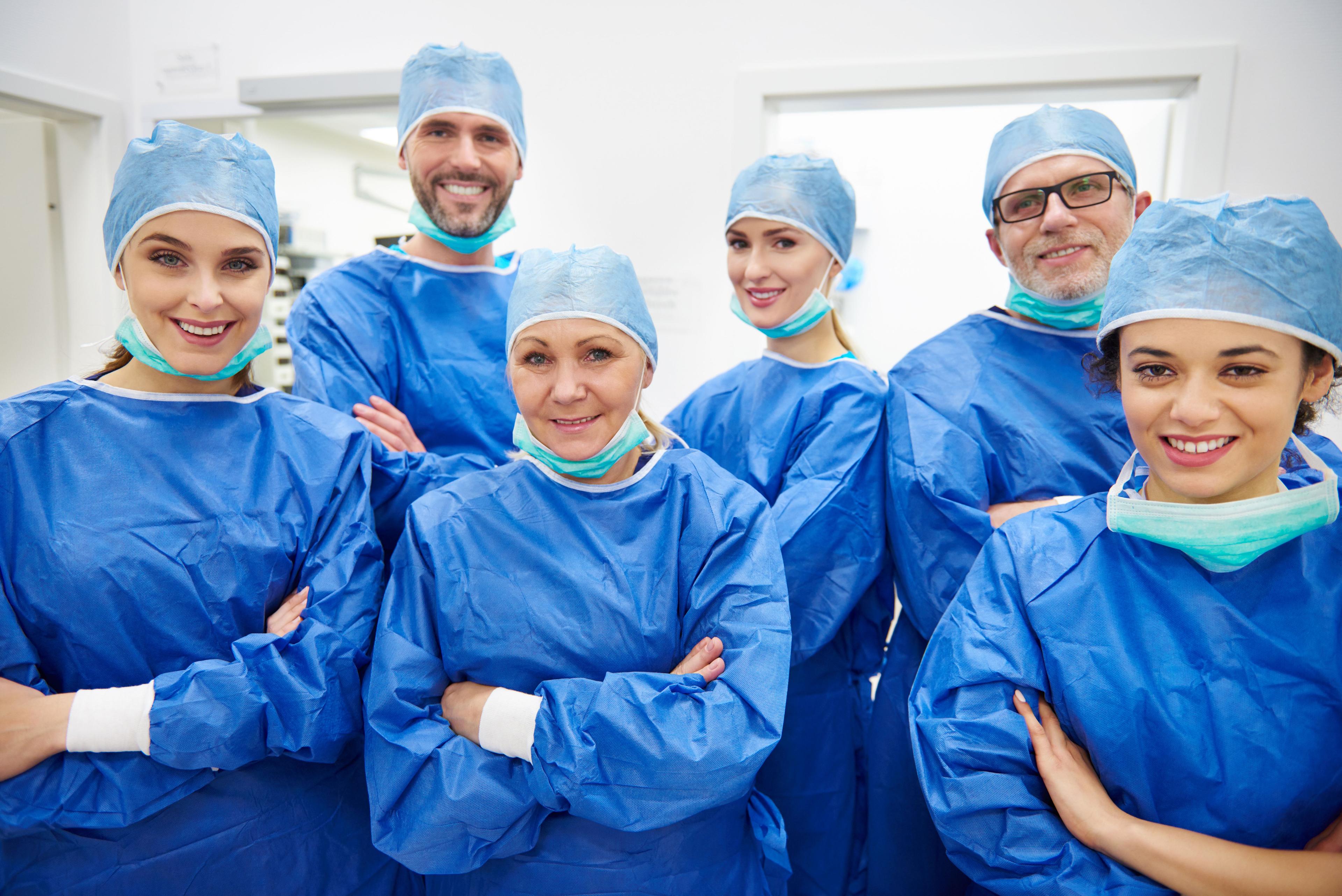 Group of six smiling spine neurosurgeons in blue scrubs and surgical caps.