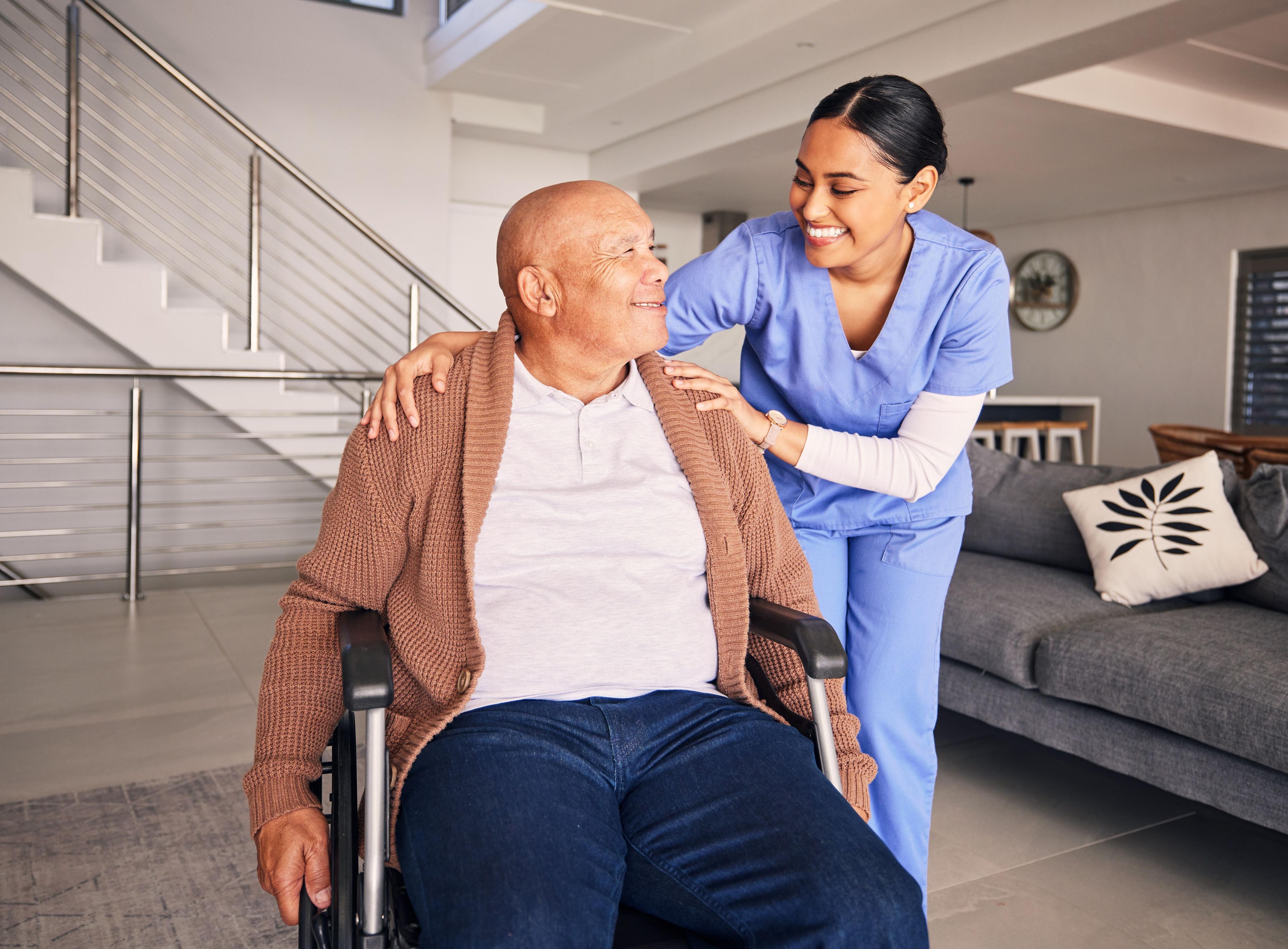 Caregiver in blue assists smiling elderly man in wheelchair.