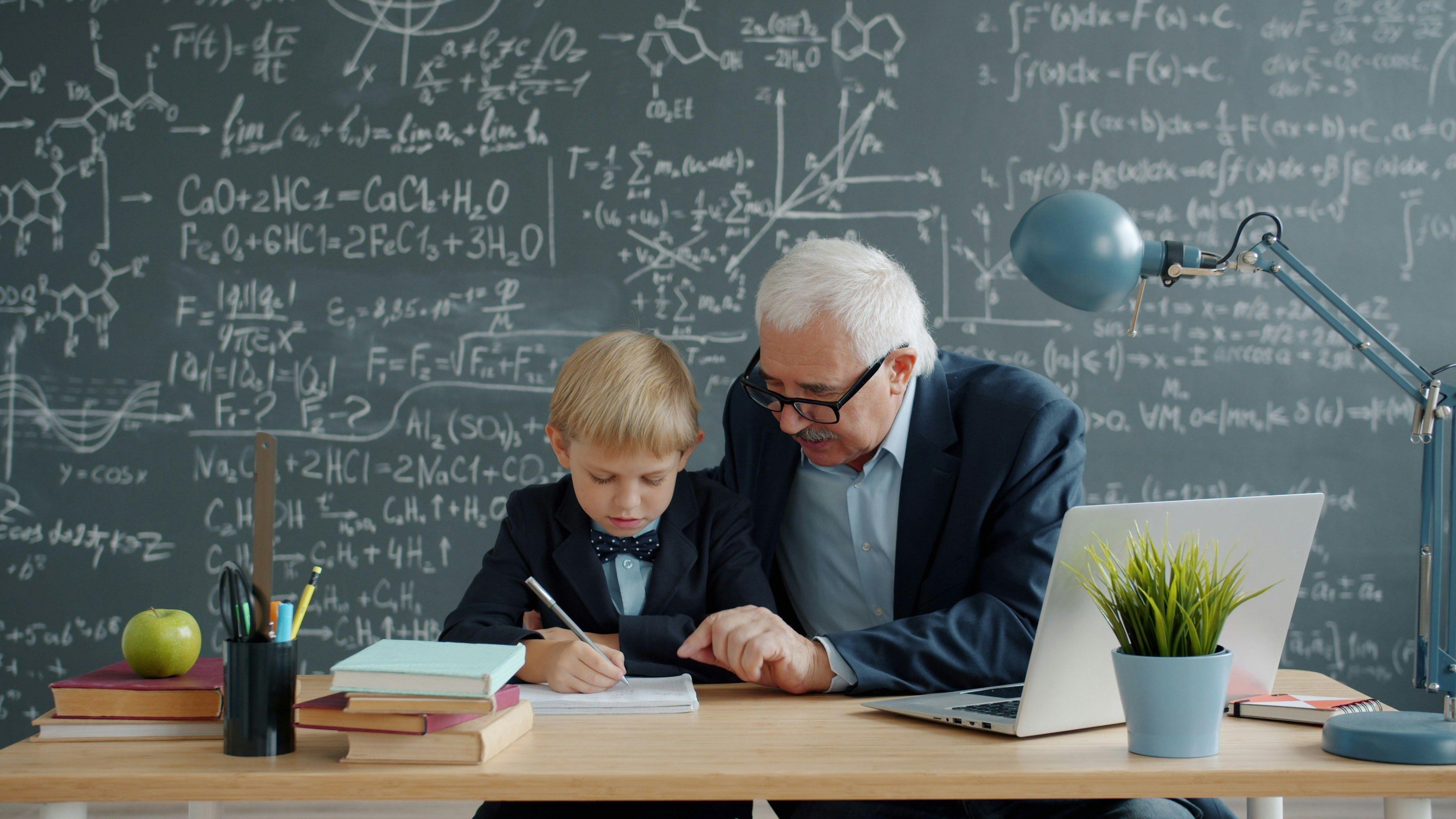 Well-dressed older man helping child with homework in front of a chalkboard with equations on it.