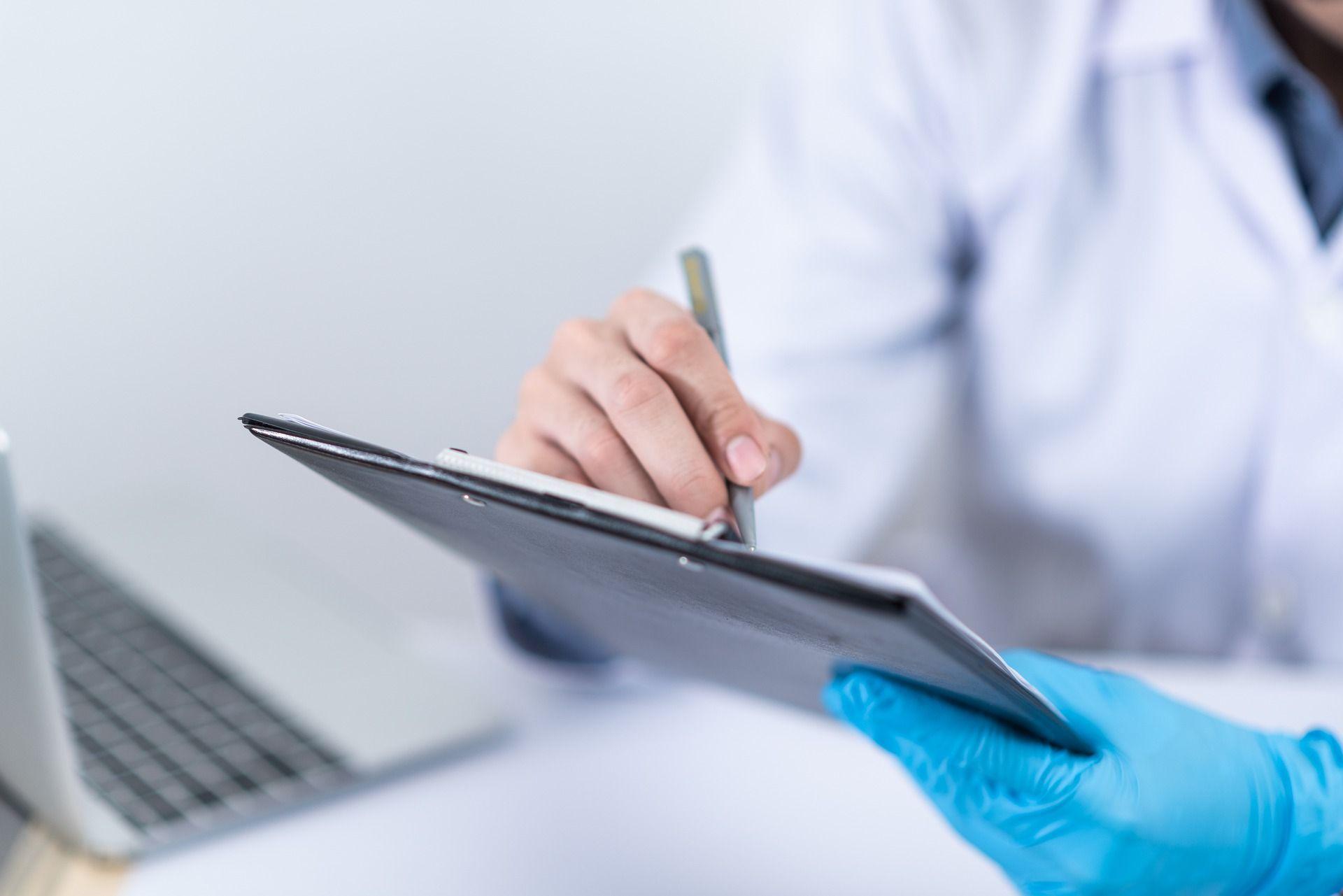 Doctor in gloves writing on a clipboard near a laptop.