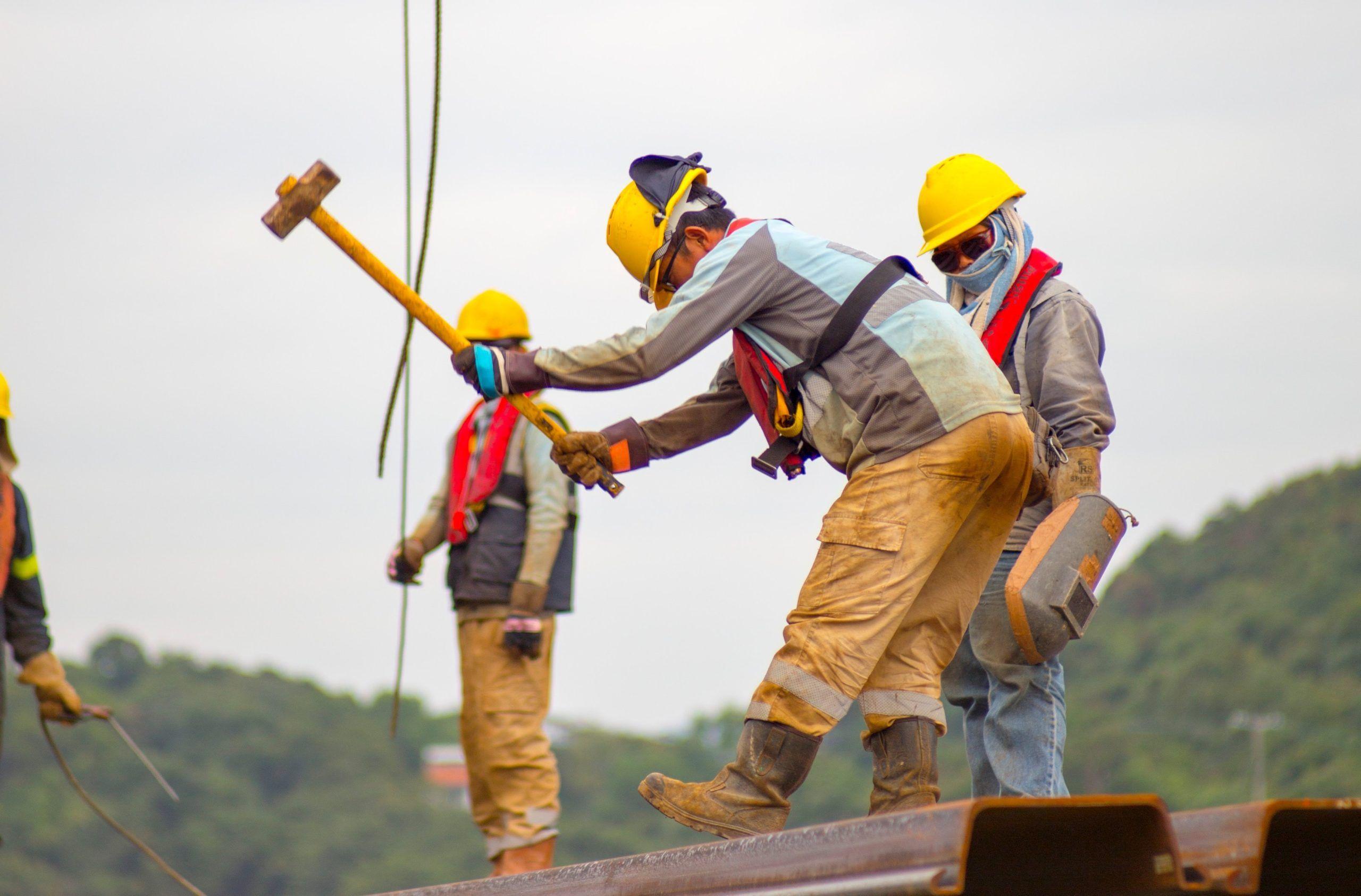 Construction workers in safety gear handling tools on a high steel beam.