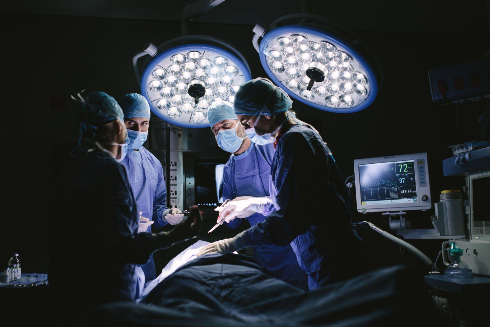 Surgeons in blue scrubs perform surgery under bright lights in an operating room.
