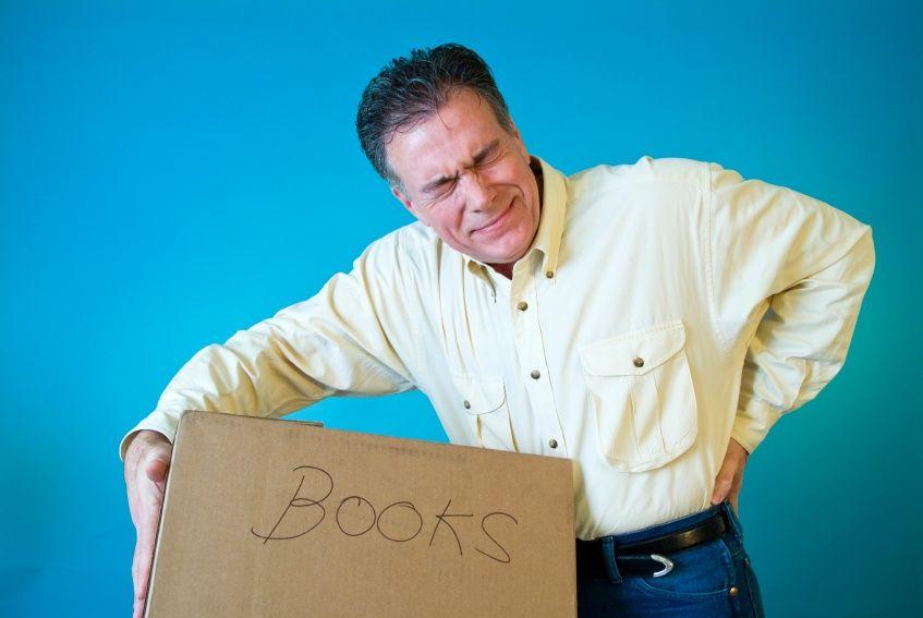 Man in cream shirt grimacing while lifting heavy cardboard box.
