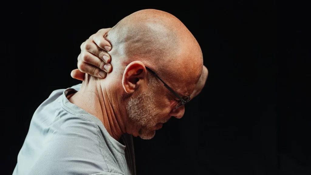 Bald man in a gray shirt grasping his head in pain or distress against a black background.
