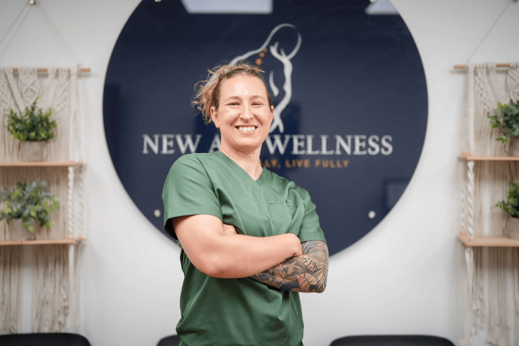 Person in green scrubs smiling in front of a "New Age Wellness" sign.