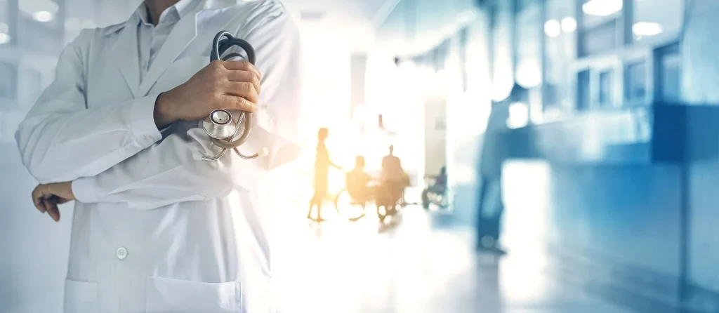 Doctor in white coat holding a stethoscope with blurred patients in background in a hospital.