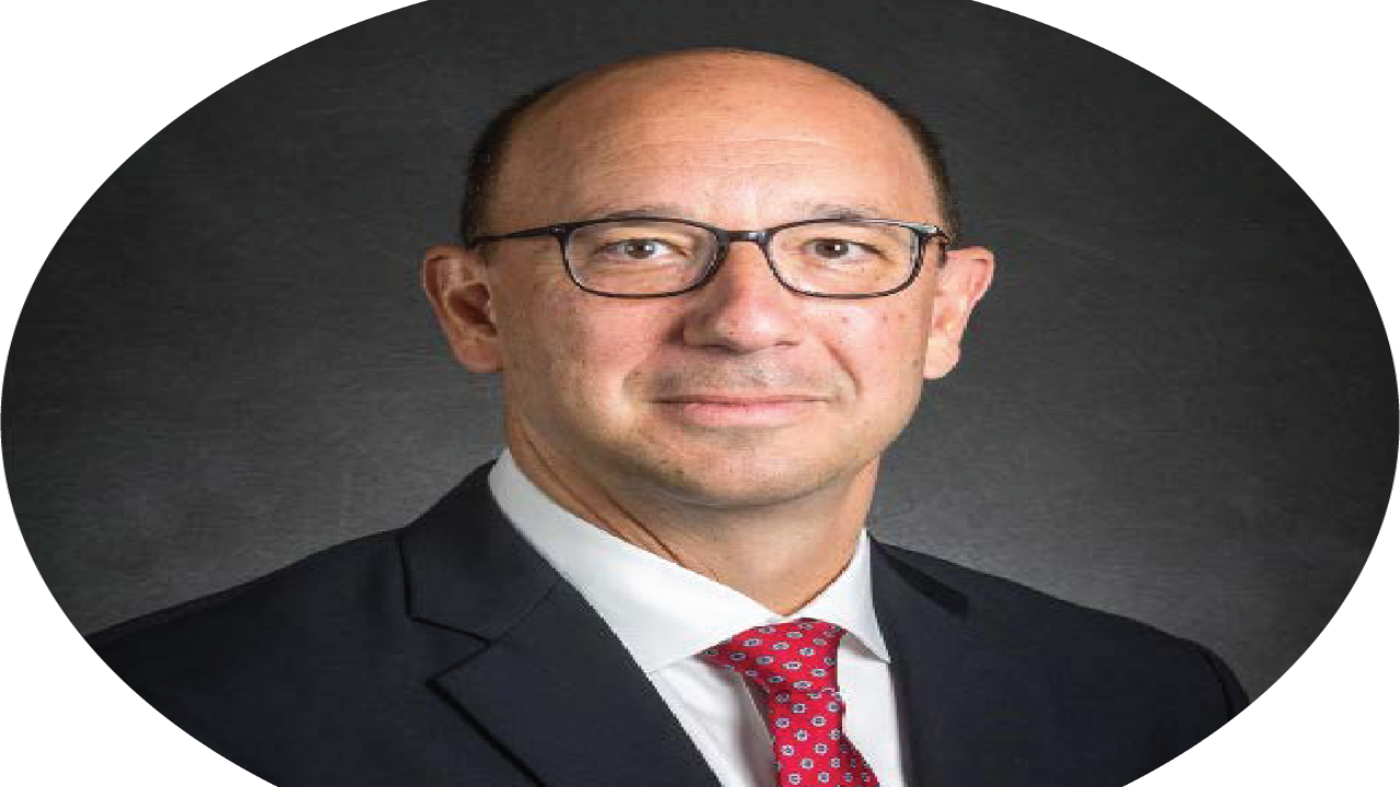A man in a suit and glasses with a neutral expression on a dark background.