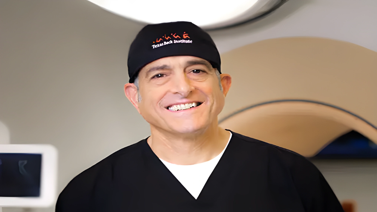 Smiling person wearing black scrubs and a cap in a medical setting.