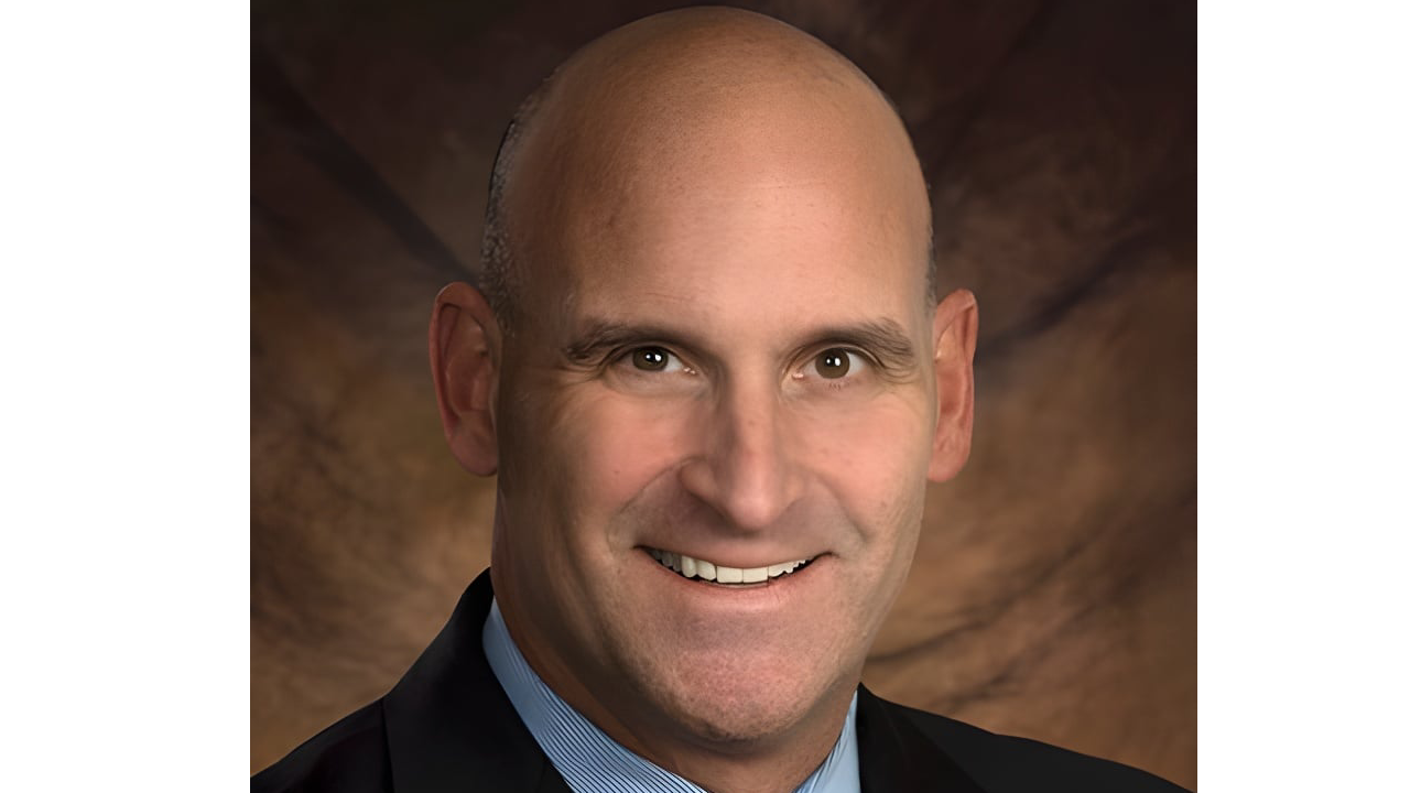 A bald man in a suit smiles against a brown, textured background.