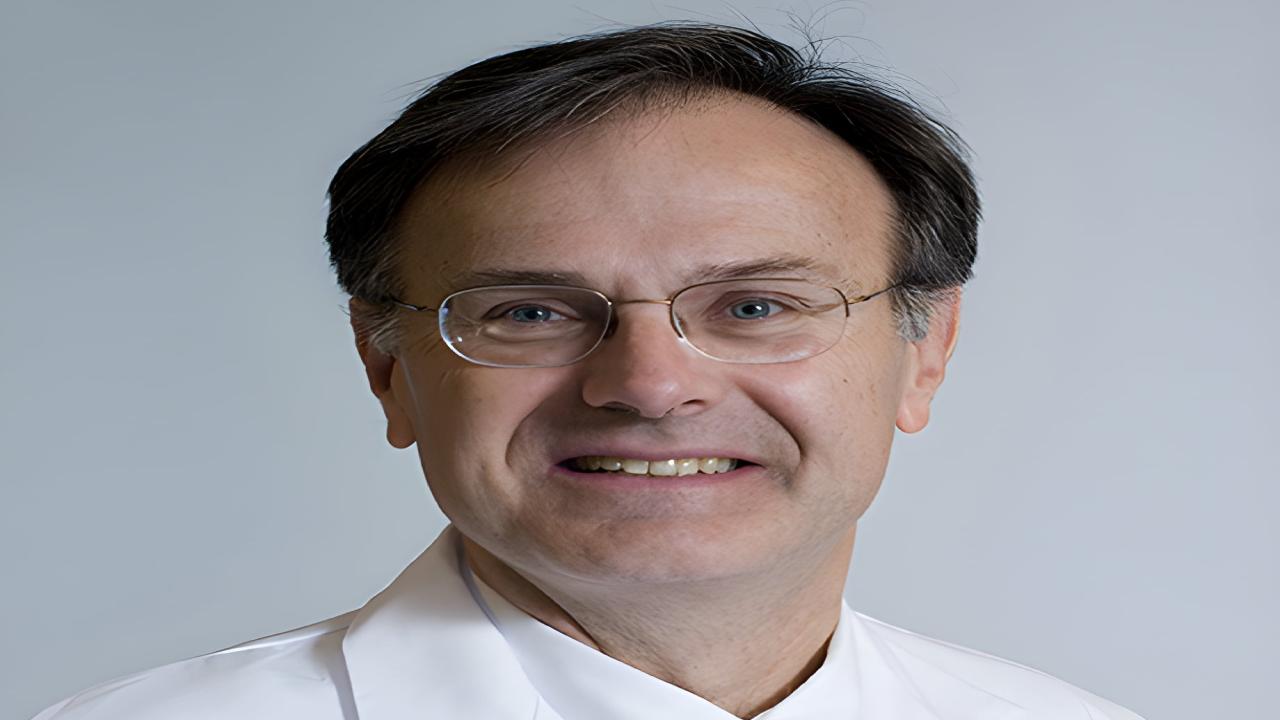 Smiling man wearing glasses and a white coat, against a plain background.