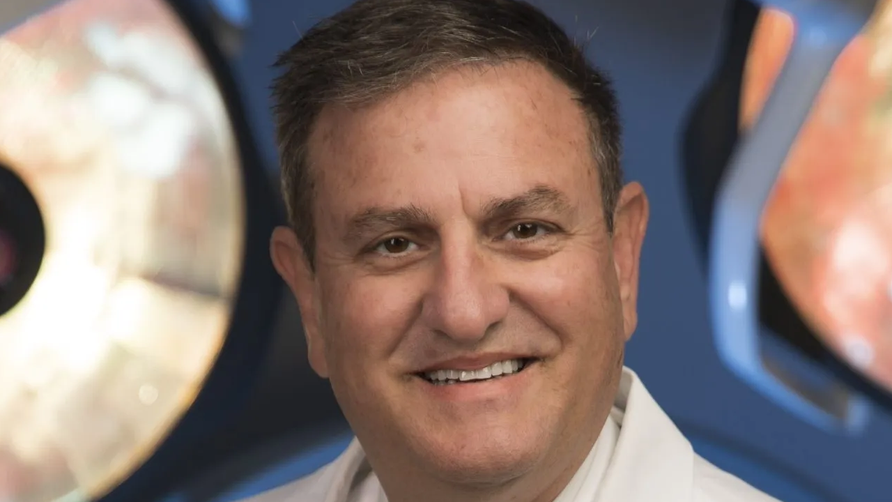 Smiling man in a white coat with a blue and orange blurred background.