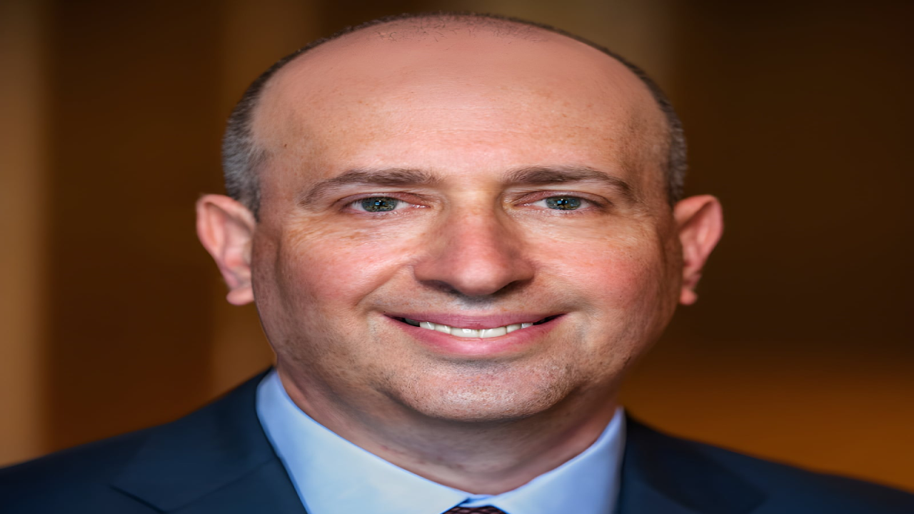 Bald man in a suit smiling against a blurred background.