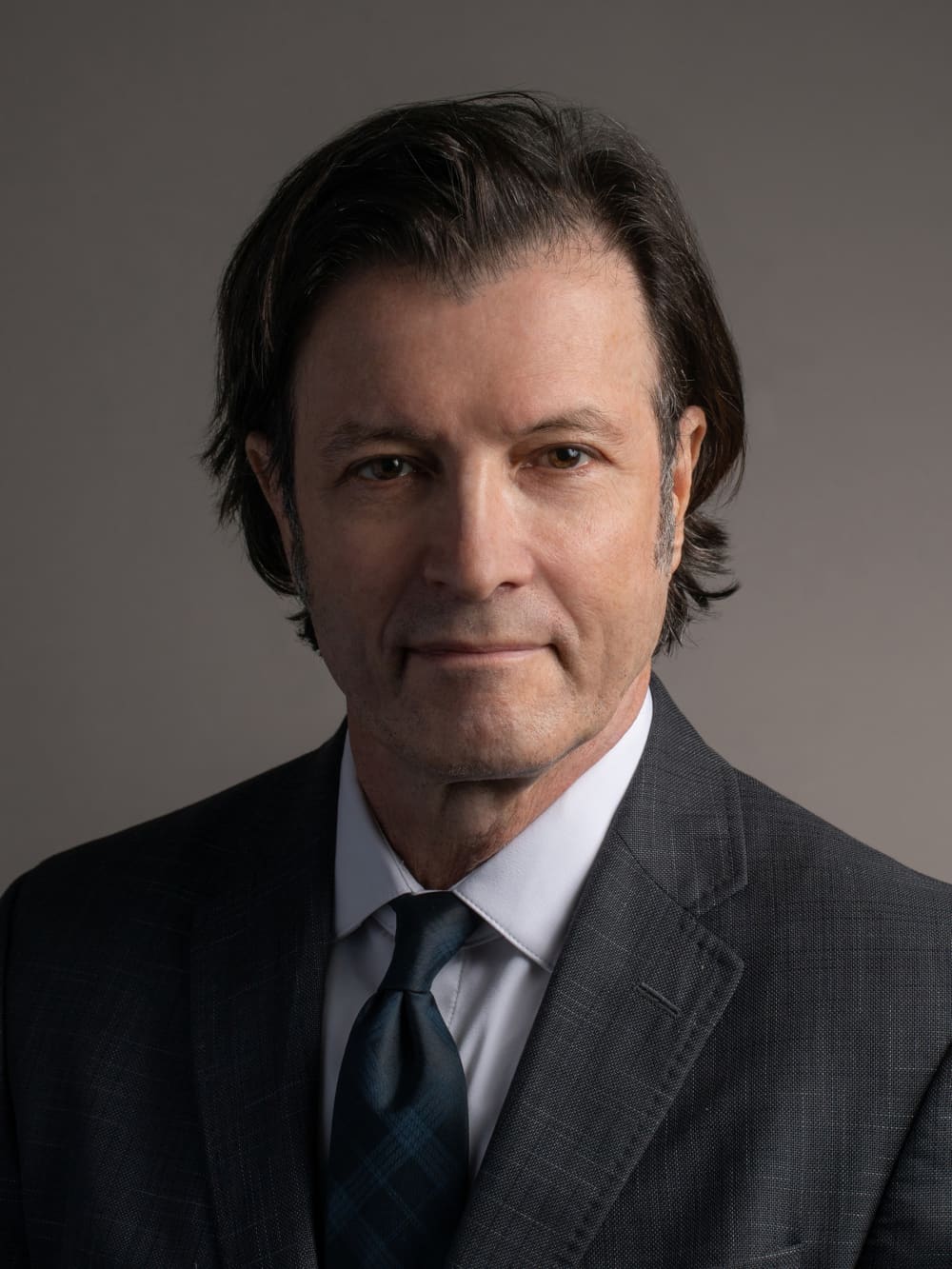 Man in a suit and tie with dark hair, against a neutral background.