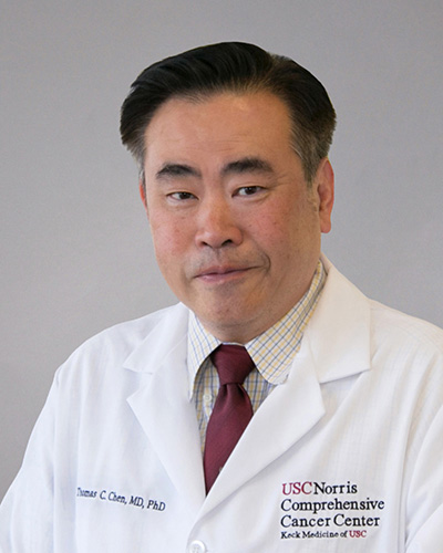 Man in a lab coat with a red tie, in front of a gray background.