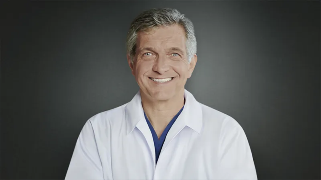 Smiling man in a white coat against a dark background.