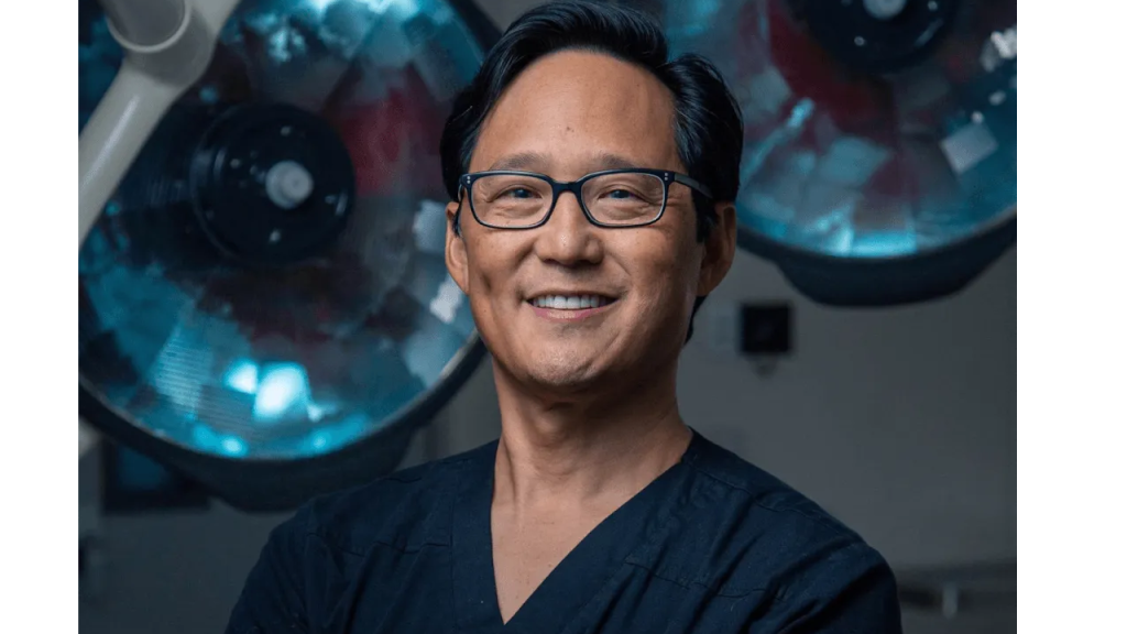 Smiling man in scrubs stands in an operating room with surgical lights in the background.