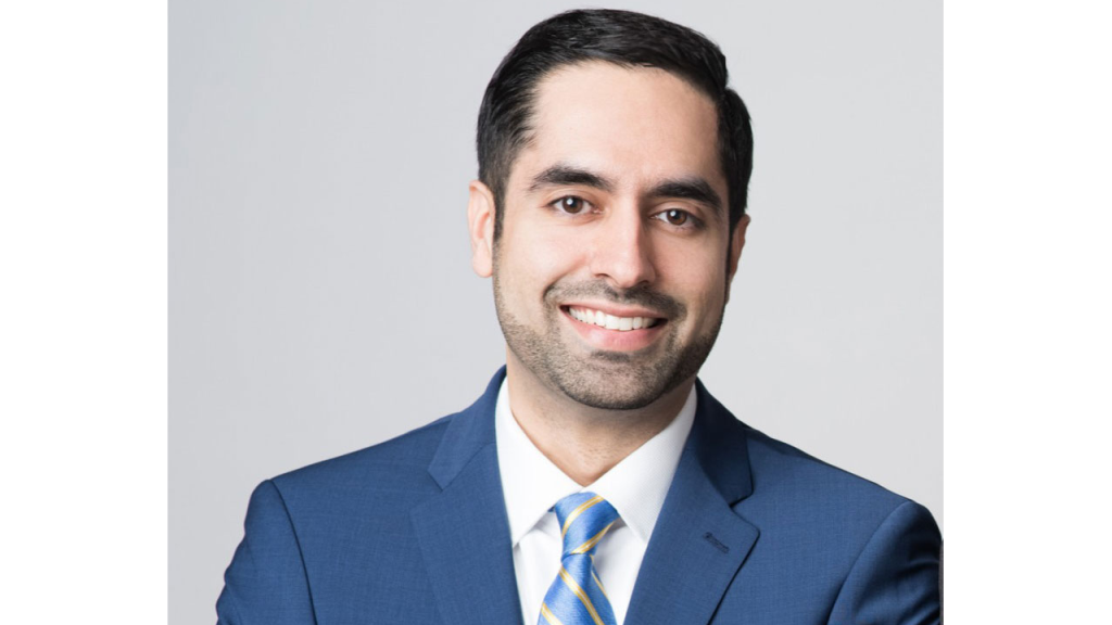 Smiling man in a blue suit with a striped tie against a gray background.