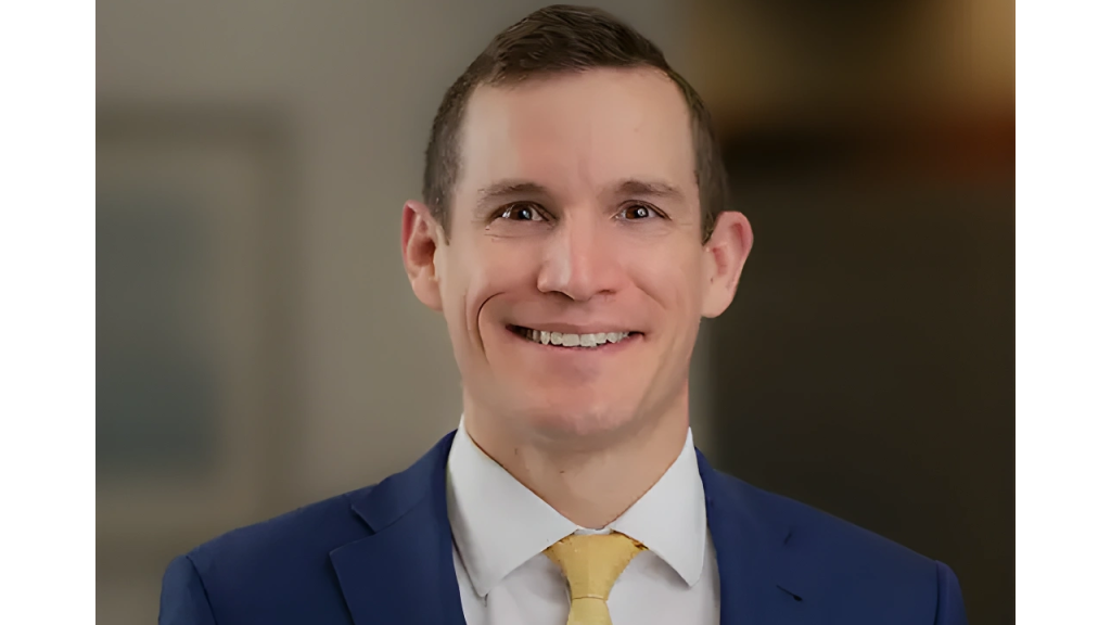 Smiling man in a blue suit and yellow tie against a blurred background.