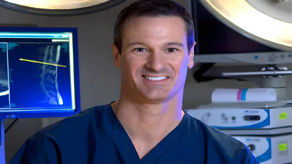 A smiling person in medical scrubs stands in a room with medical equipment and a screen displaying an X-ray.