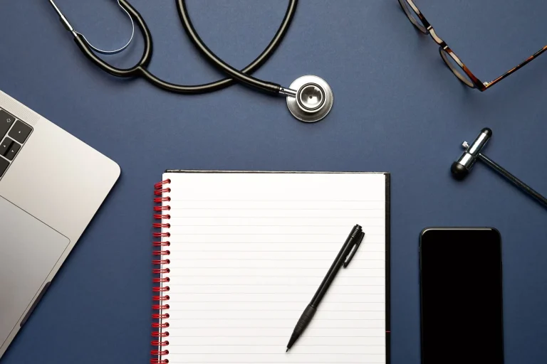 Office desk with a laptop, stethoscope, notepad, pen, glasses, reflex hammer, and smartphone on a blue surface.