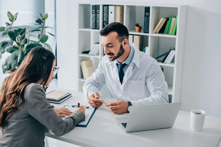 A doctor consults with a patient in an office setting, with a laptop and clipboard on the desk.