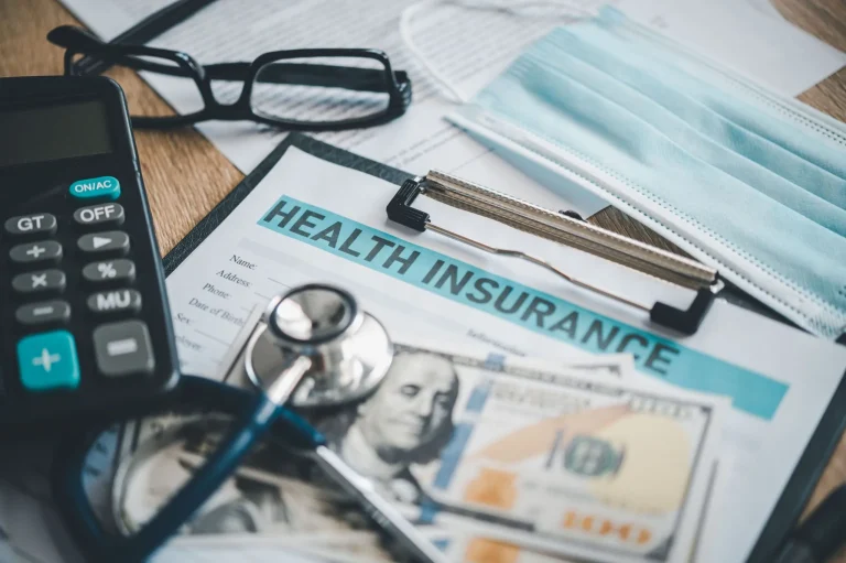 Health insurance documents, a stethoscope, mask, calculator, glasses, and cash on a desk.