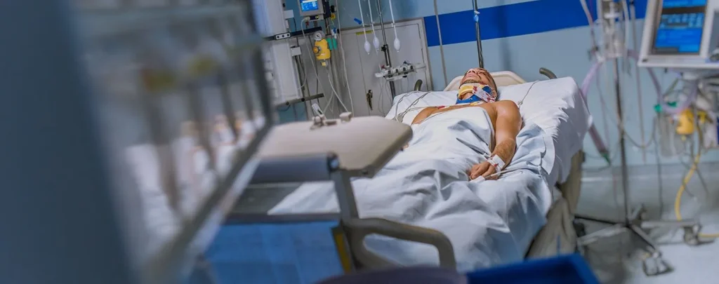 A person with a neck brace lies in a hospital bed surrounded by medical equipment.