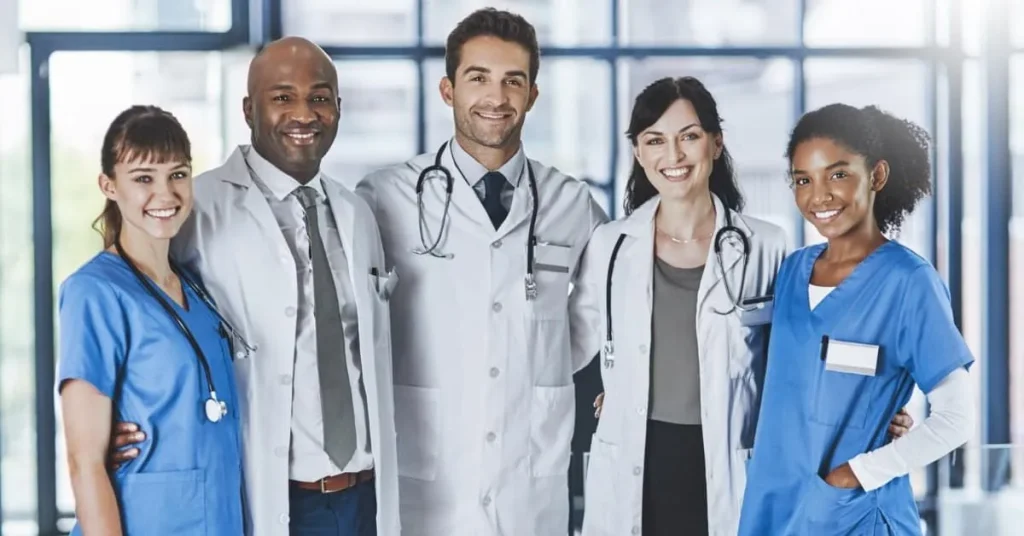 Group of five smiling medical professionals in scrubs and lab coats stand together.