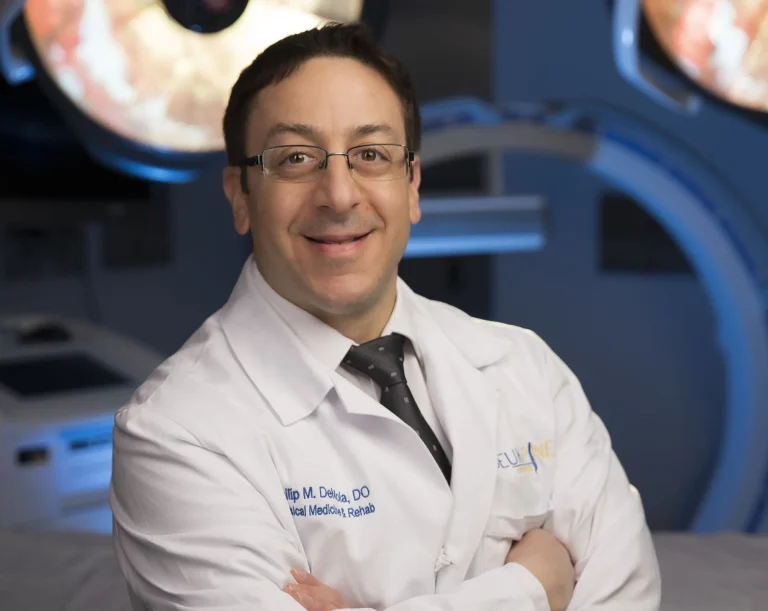 Smiling person in a white medical coat and glasses, standing in a medical setting.