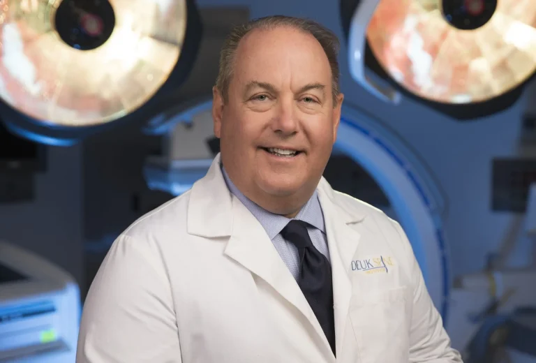 Smiling man in a white coat with surgical lights in the background.
