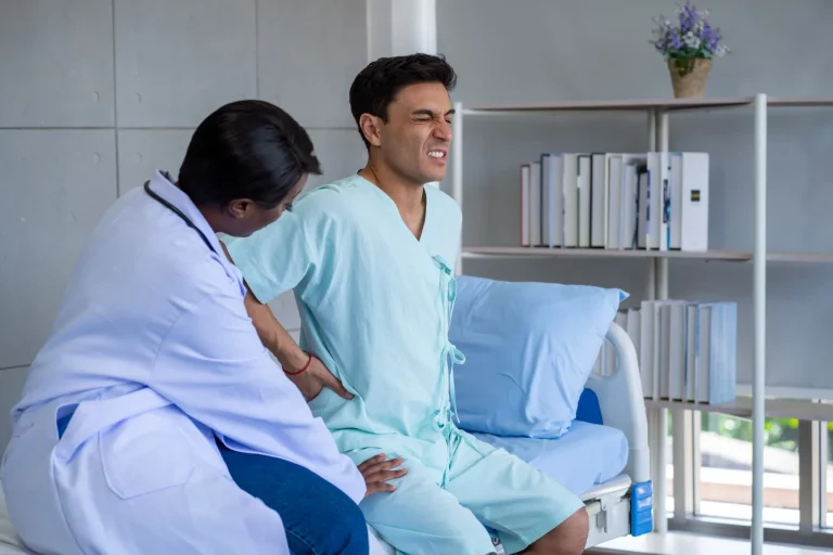 A doctor examines a man in a hospital gown, grimacing in pain while sitting on a bed.