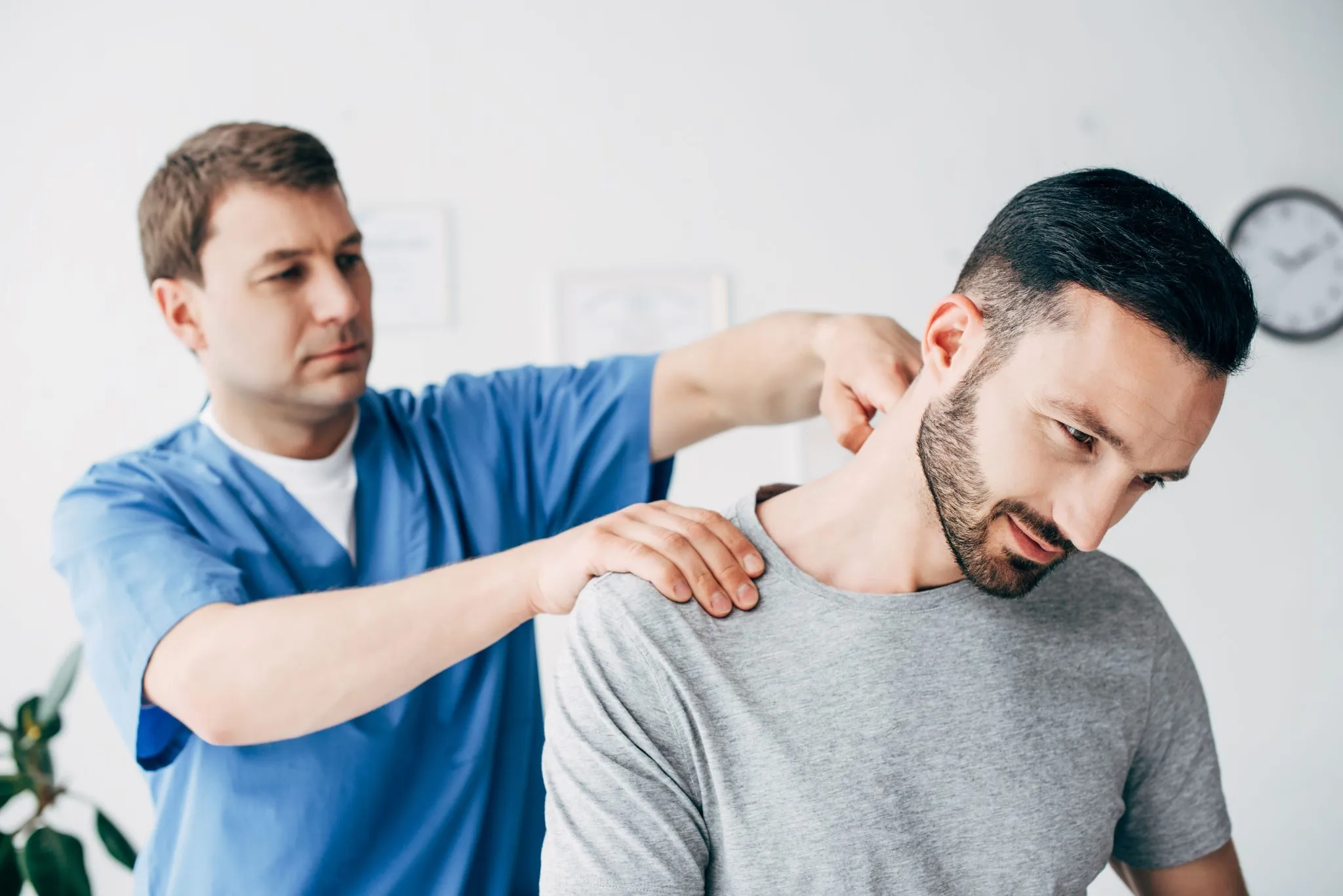 A male healthcare professional is examining a male patient's shoulder and neck area in a clinical setting, attempting to accurately diagnose the cause of the patient's pain.