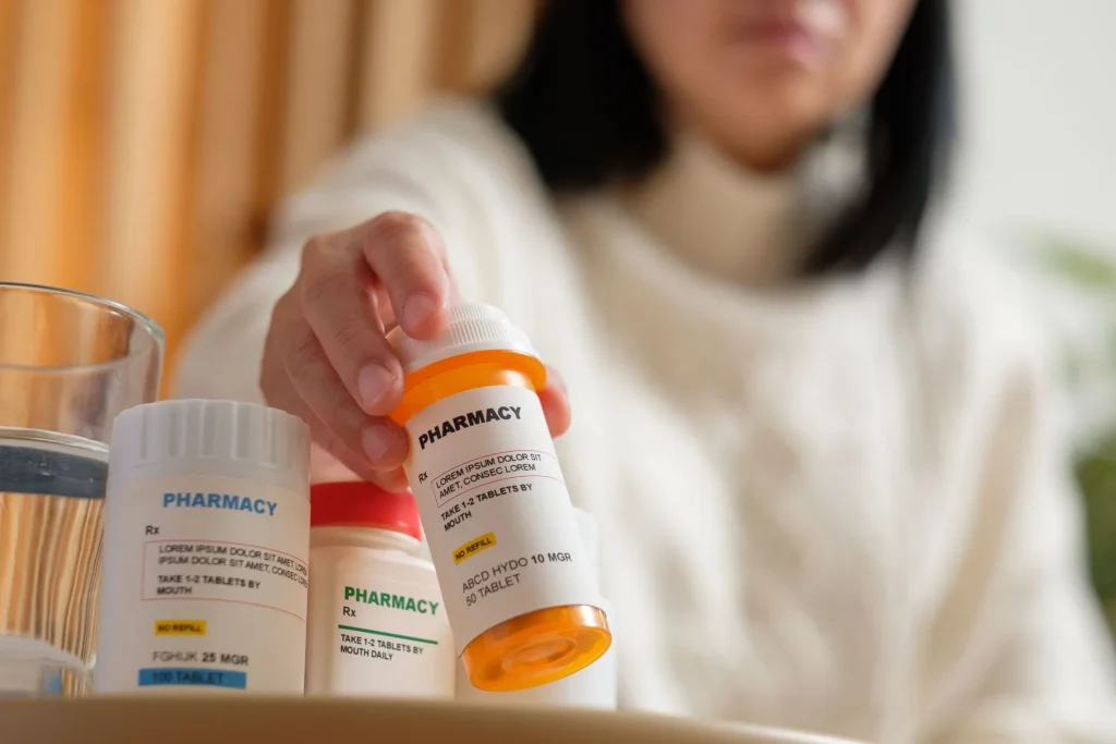 Person holding a prescription pill bottle near other medication bottles and a glass of water.