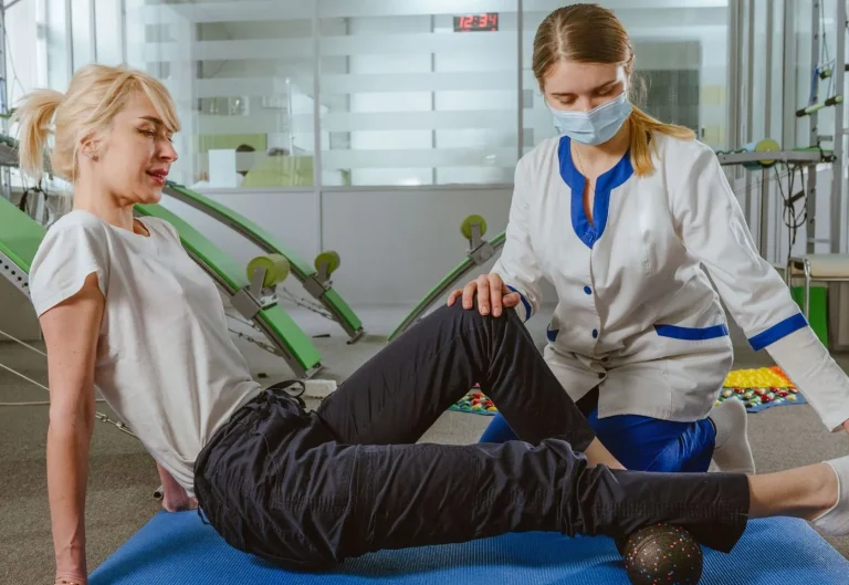 A physiotherapist assists a woman stretching her leg on a blue mat in a clinic.