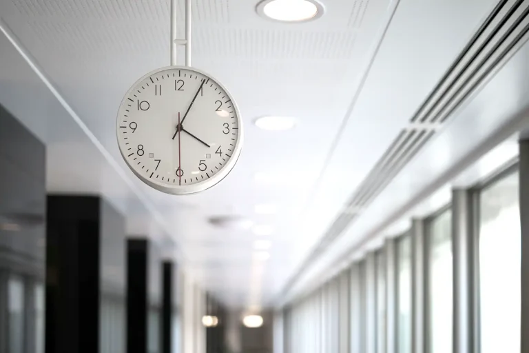 A round white clock hangs from the ceiling in a bright corridor.