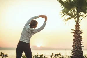 Person stretches near a palm tree at sunrise by the water.