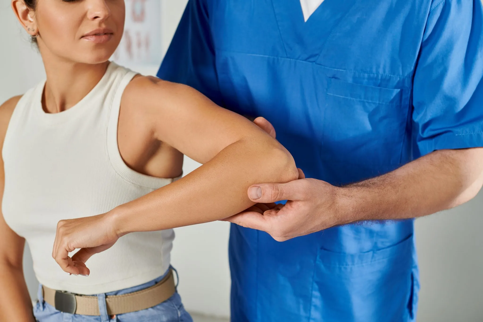 A physical therapist in blue scrubs examining a woman's elbow, demonstrating how physical therapy treats various musculoskeletal issues.