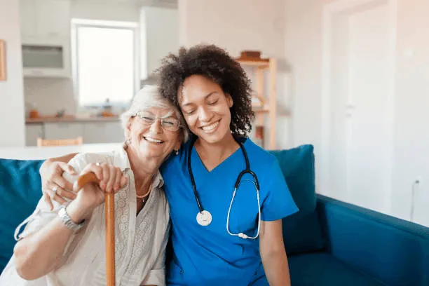 A nurse in blue scrubs sits smiling with an elderly woman holding a cane.