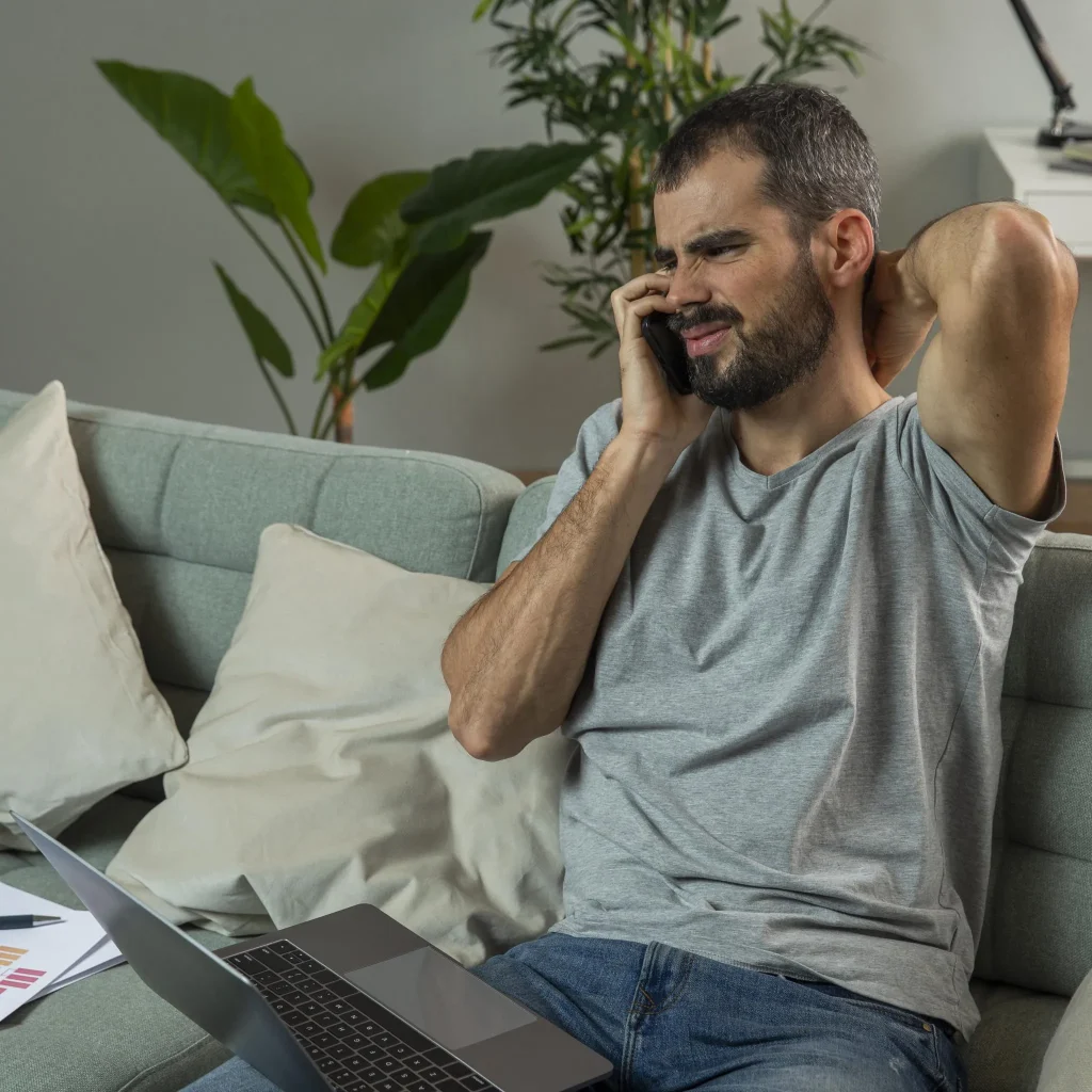 Man on a sofa talking on a phone, looking stressed, with a laptop and papers nearby.