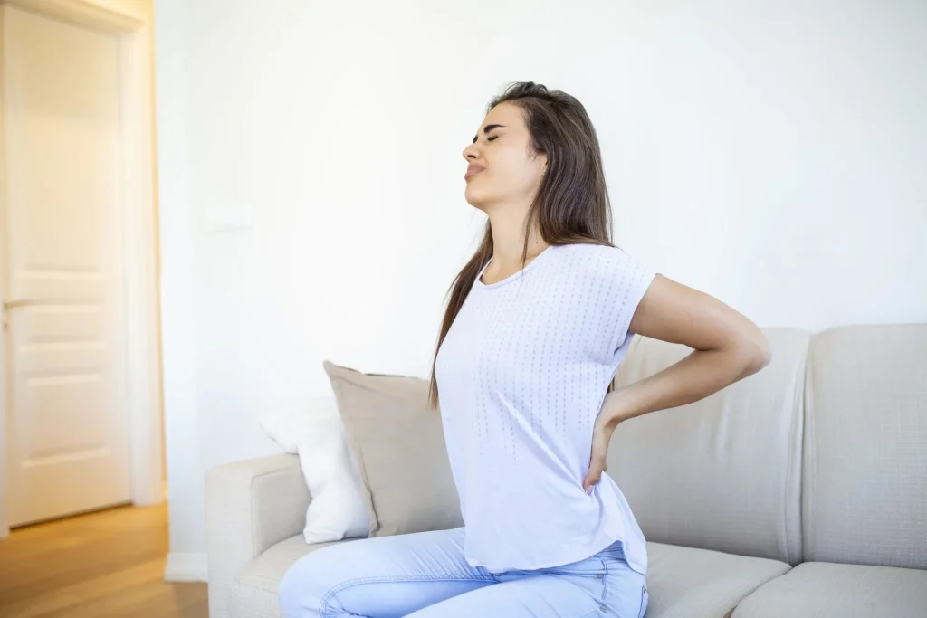A woman sitting on a sofa, holding her lower back in pain.