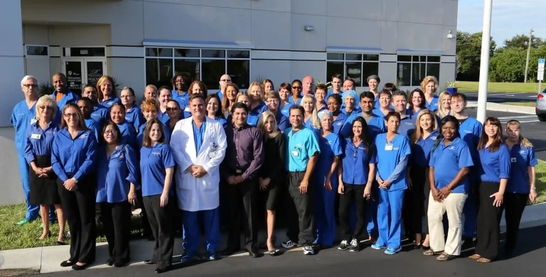 A large group of people in blue uniforms and a doctor in a white coat pose outside a building.