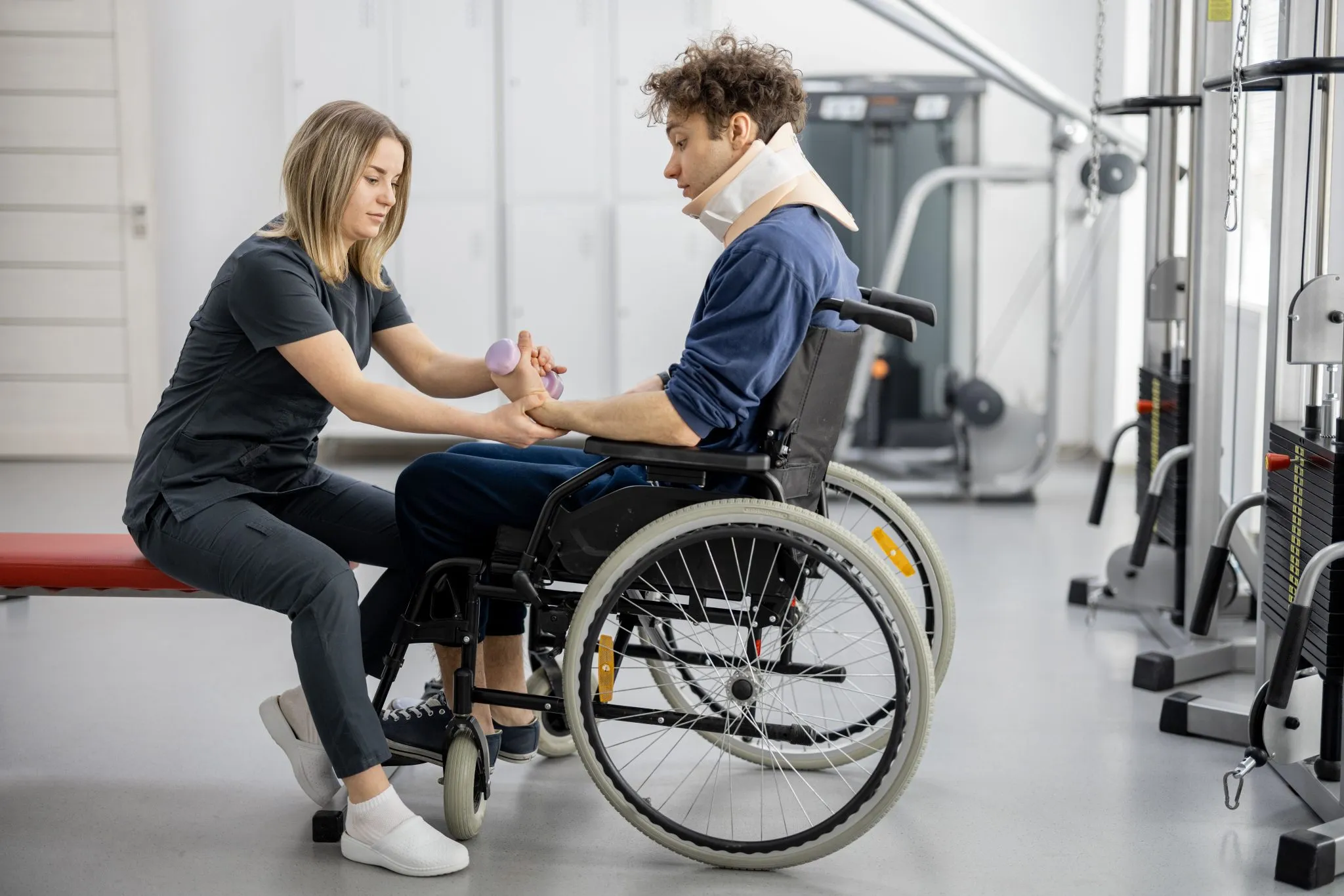 A man in a wheelchair with a neck brace receives physical therapy from a female therapist in a modern rehabilitation center, illustrating a patient potentially dealing with complications such as paralysis after an epidural steroid injection.