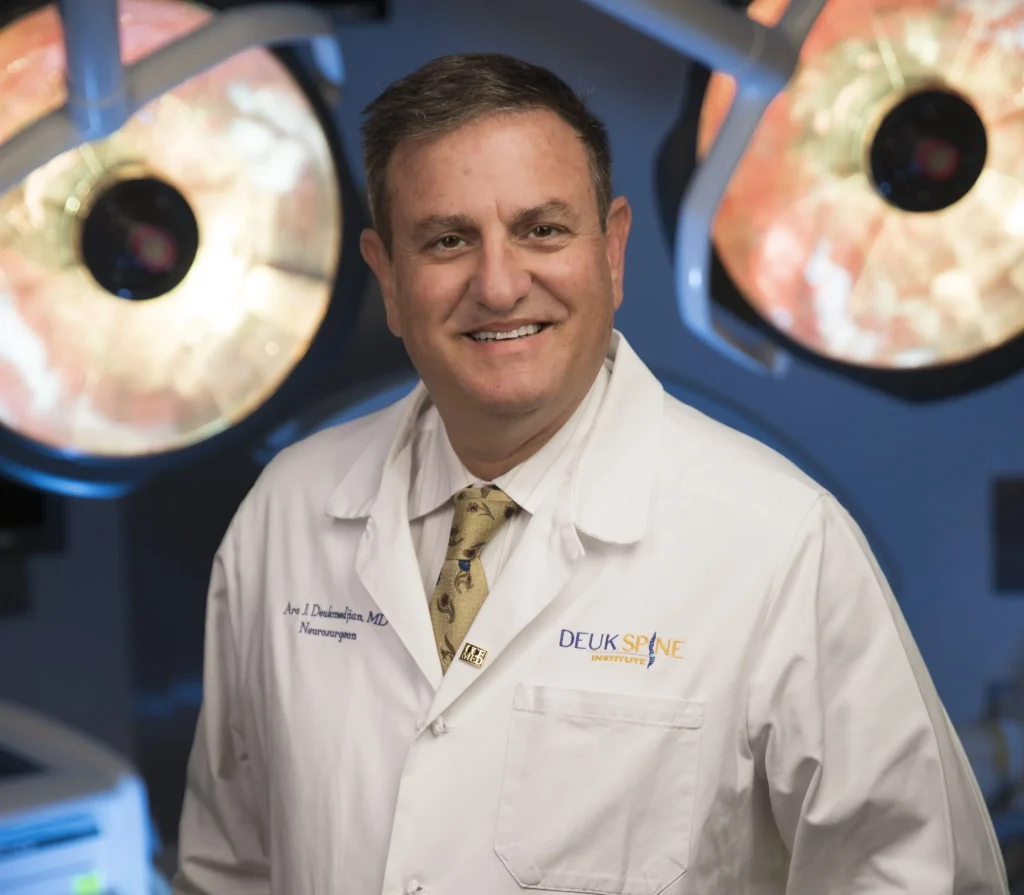 A doctor in a white coat smiles in an operating room with overhead surgical lights.