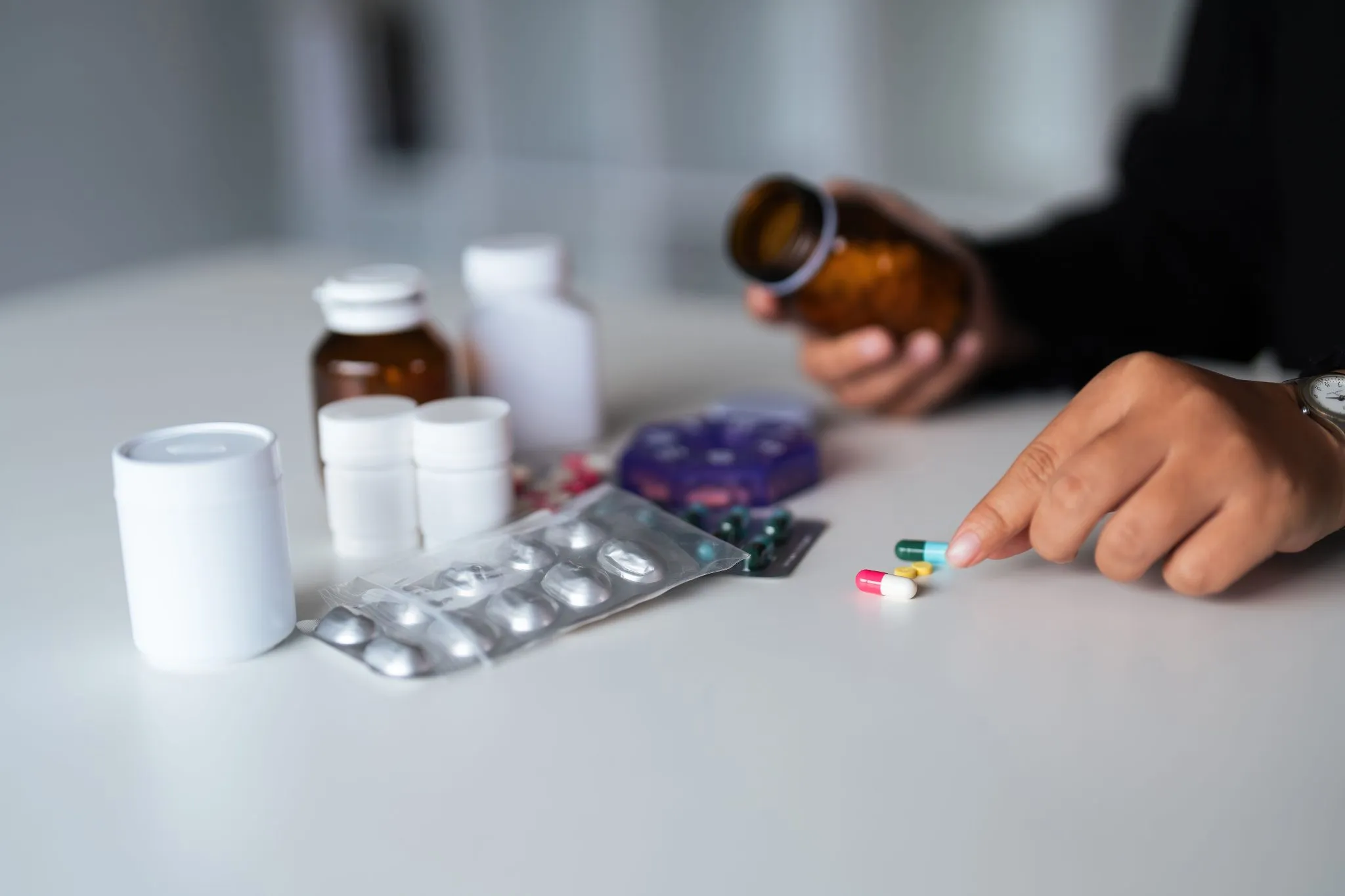 A person's hand selecting pain relief pills from various medicine bottles and packages on a table.