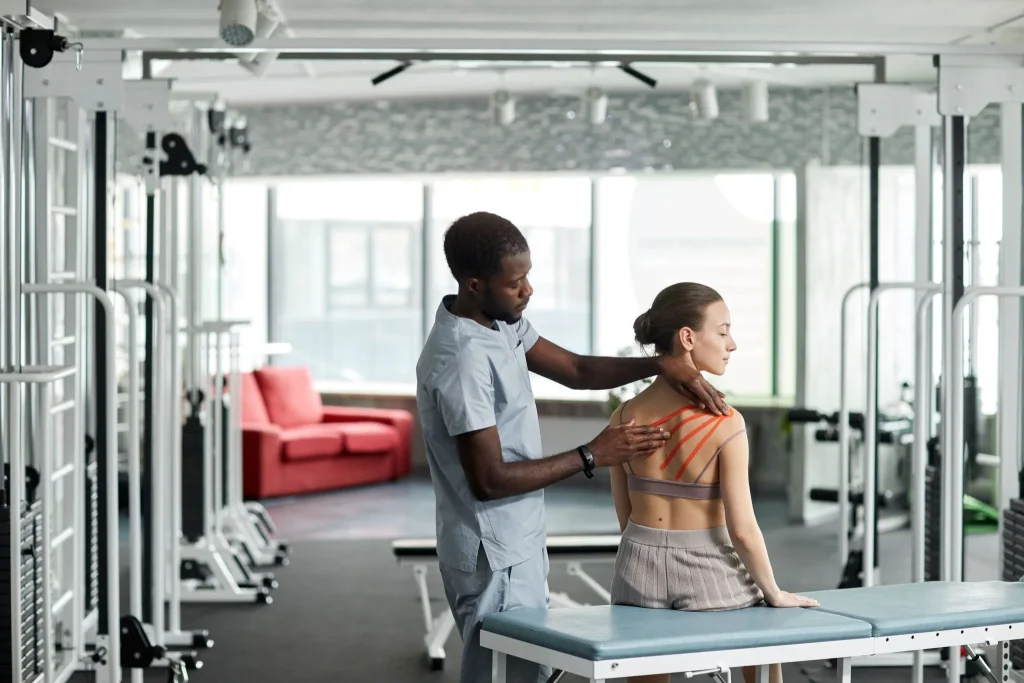 A therapist applies kinesiology tape to a woman's shoulder in a gym setting.