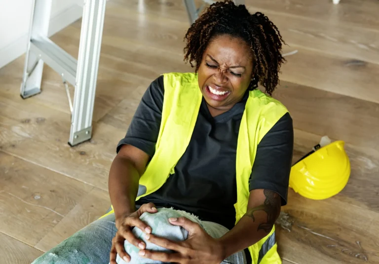 Person in a yellow vest sitting on the floor in pain, holding their knee, with a hard hat nearby.