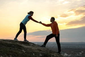 Two people hiking on a hill, one helping the other up at sunset.