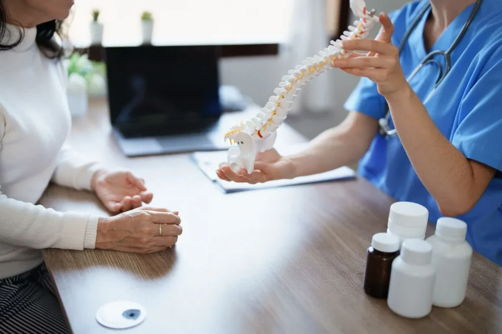 A doctor shows a spine model to a patient at a desk with medication bottles.