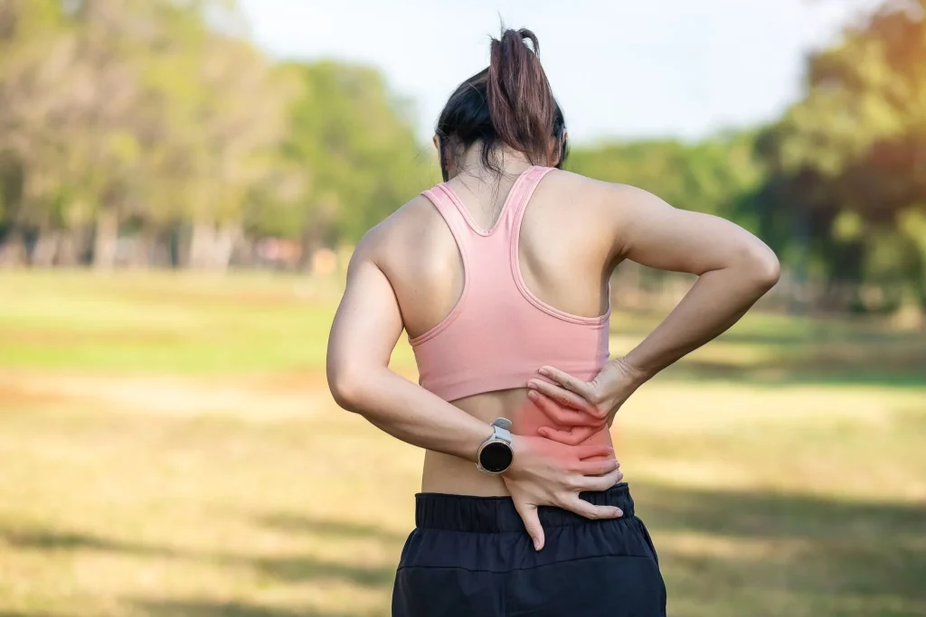 A woman in athletic wear holding her lower back in discomfort outdoors.